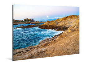 Acrylic photo print of a rocky coastline with blue ocean waves and distant buildings at sunset