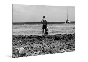 Black and white acrylic photo print of two people on rocky beach with sailboat in background