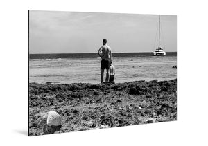Black and white aluminum photo print of two men on a rocky beach with a sailboat in the distance