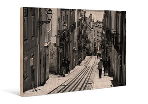 Black and white print of a narrow city street with tram tracks and pedestrians on wood panel