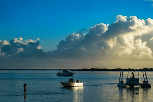 Serene coastal scene at sunset with boats on calm water, dramatic clouds, and a child wading.