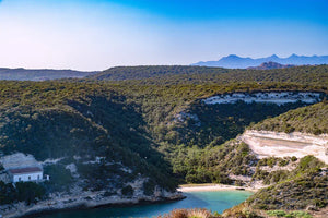 Coastal landscape with rugged cliffs, turquoise bay, green hills, and a white house