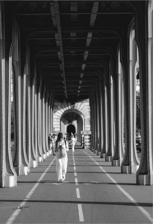 Photo noir et blanc de Paris sous un viaduc avec forte perspective, colonnes métalliques et silhouettes marchant vers une arche lumineuse