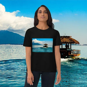 Woman in black t-shirt with tropical boat and ocean print, standing by serene blue water.