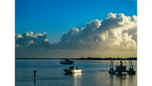 Child standing in shallow water at sunset with boats and dramatic clouds over the sea
