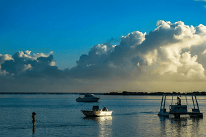 Serene seascape at sunset with boats, dramatic clouds, and a person standing in shallow water.