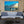 Modern living room with light blue sofa, potted plant, and wall art of boats on tranquil water at sunset.