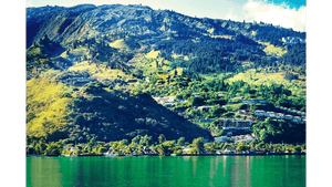 Colorful landscape photo of lush green mountains and houses by a turquoise lake under blue sky