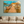 Wall art of a tropical pier with boats and thatched-roof hut, displayed above beige sofa