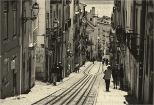 Sepia photo of people walking along a narrow European street with tram tracks, old buildings and lanterns.