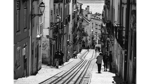 Black and white photo of a steep European street with tram tracks, old buildings, and pedestrians.