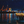 Nighttime harbor with illuminated dockside restaurant, cranes, and water reflections under stars