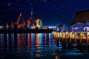 Vibrant port at night with dockside restaurant, illuminated cranes, and starry sky reflection.