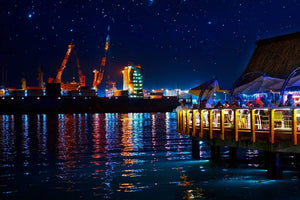 Vibrant port city night scene with illuminated waterfront restaurant, cranes, and starry sky.