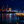 Nighttime harbor scene with illuminated dockside restaurant, cranes, and starry sky reflection on water