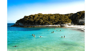 People swimming in turquoise water near a sandy beach and tree-covered rocky island