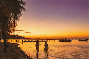 Tropical beach sunset with palm trees, people on the shore, boats, and a wooden pier