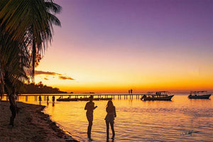 People on tropical beach at sunset with palm trees, pier, and boats on calm ocean water.