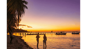 Tropical beach sunset with palm trees, people by the shore, boats and a pier over the ocean