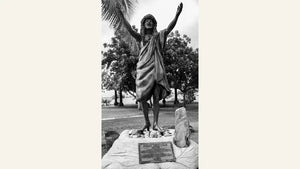Black and white photo of Polynesian statue with outstretched arms in a tropical park setting.