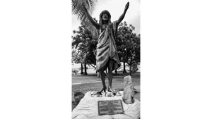 Black and white photograph of a Polynesian statue with arms raised, outdoors among trees.