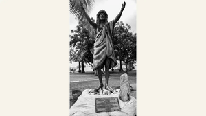 Black and white photo of Polynesian statue with arms raised, outdoor setting, palm tree nearby.