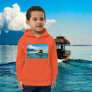 Child in orange eco-friendly hoodie with tropical ocean and hut print, standing by water