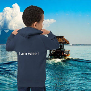 Child wearing navy ecological hoodie with 'I am wise!' text, standing by tropical water and boat.