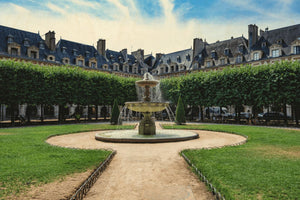 Image représentant la fontaine, le jardin et les façades historiques de la place des Vosges à Paris sous un ciel bleu