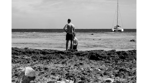 Black and white photo of two men on a rocky beach looking at a sailboat on the horizon