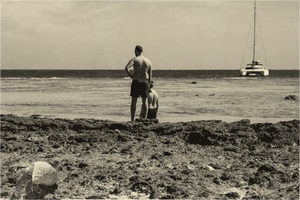 Two people standing on a rocky beach facing the sea with a distant sailboat on the horizon.