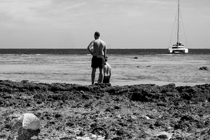 Black and white fine art photo of two men on a rocky beach facing the ocean with a sailboat in the distance and a large shell in the foreground.