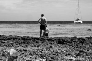 Black and white photo of two men on rocky beach, facing ocean with sailboat in background