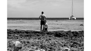 Black and white photo of two men on a rocky beach facing the ocean with a sailboat in the distance.